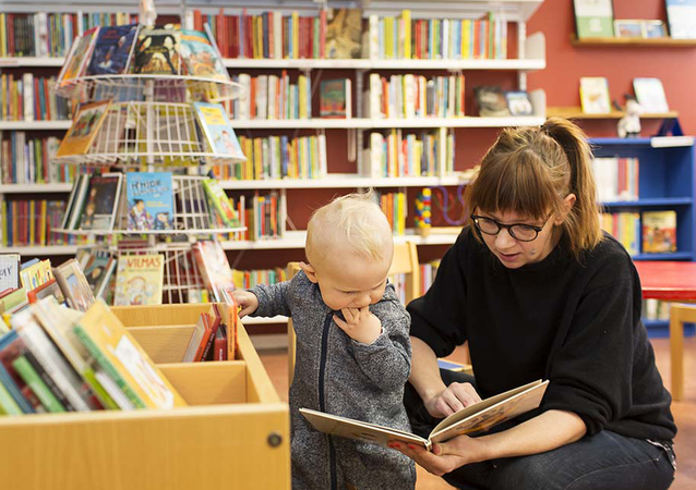 En mamma läser ur en bok för en ettåring bland böcker på ett bibliotek