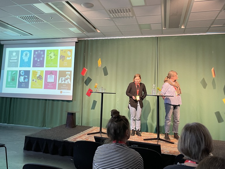 Emma och Hanna på Svedala bibliotek på scenen berättar om projektet Hej Dewey och visar skyltning över de olika avdelningarna.